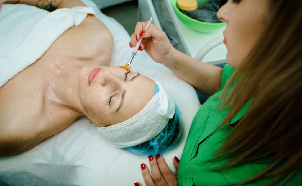 Professional Cosmetology. Beautician At Spa Salon Applying Clay Face Mask To Young Woman, Relaxed Female Enjoying Wellness And Skin Care Treatments.