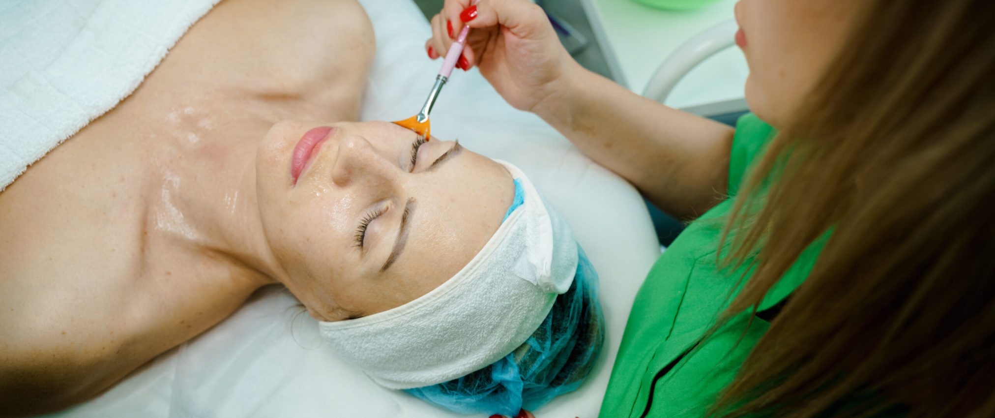 Professional Cosmetology. Beautician At Spa Salon Applying Clay Face Mask To Young Woman, Relaxed Female Enjoying Wellness And Skin Care Treatments.