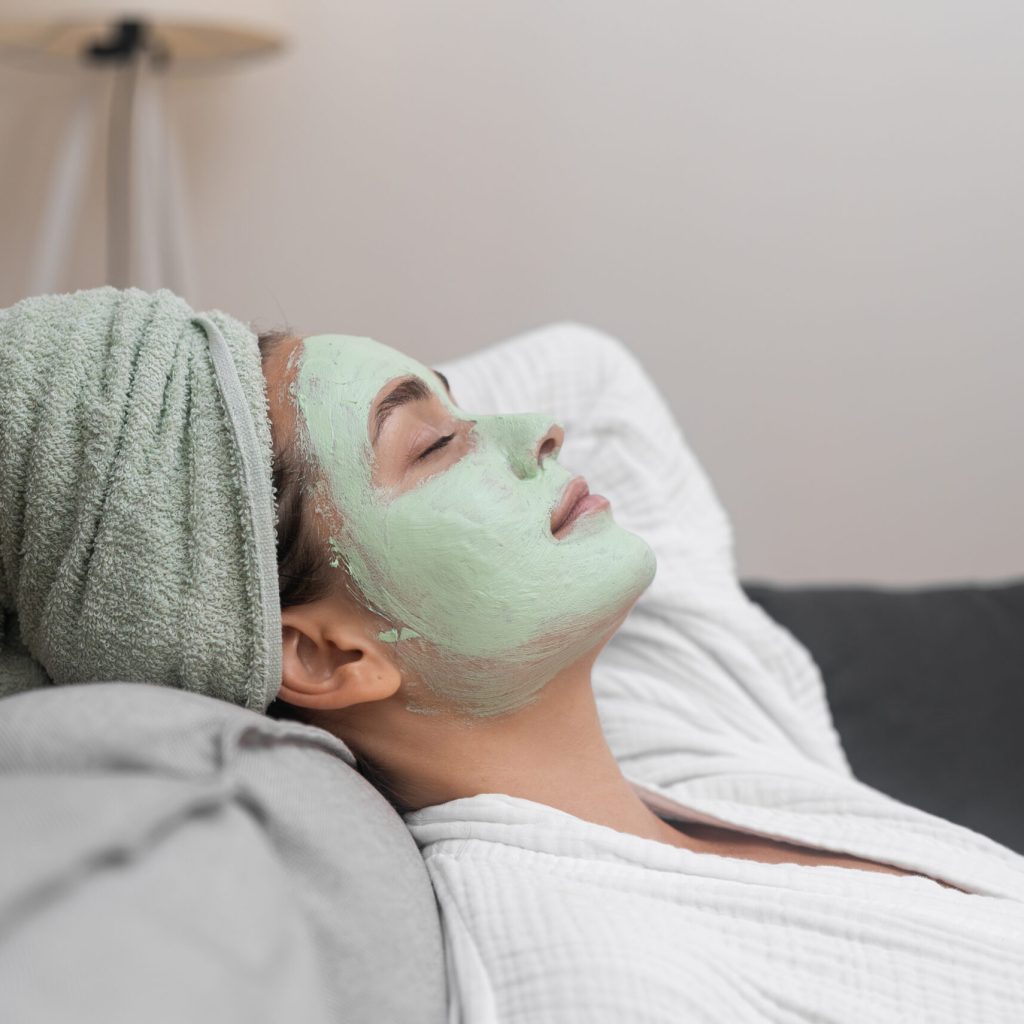 Relaxed young woman with peeling clay mask on face lying on comfortable sofa pretty lady in bathrobe resting during spa procedures with natural cosmetics